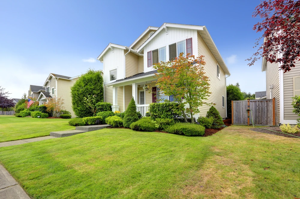 Fresh lawn mowing creates a smooth green yard around a cream two-story home with sculpted shrubs and a red-leafed tree.